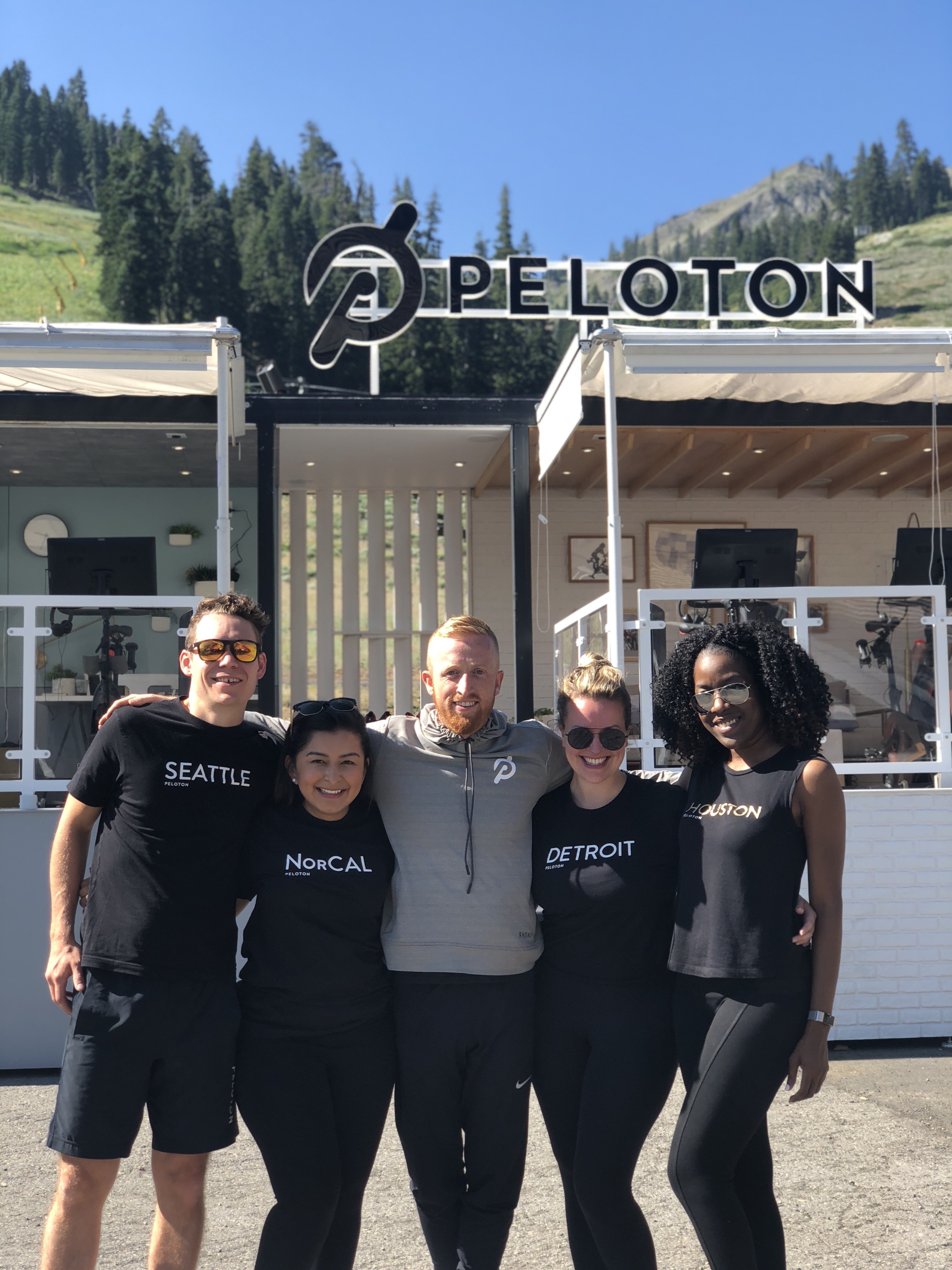 Five Peloton employees with shirts labeled with different U.S. regions they represent, huddle together outside of a mobile Peloton showroom in Aspen on a sunny summer day.