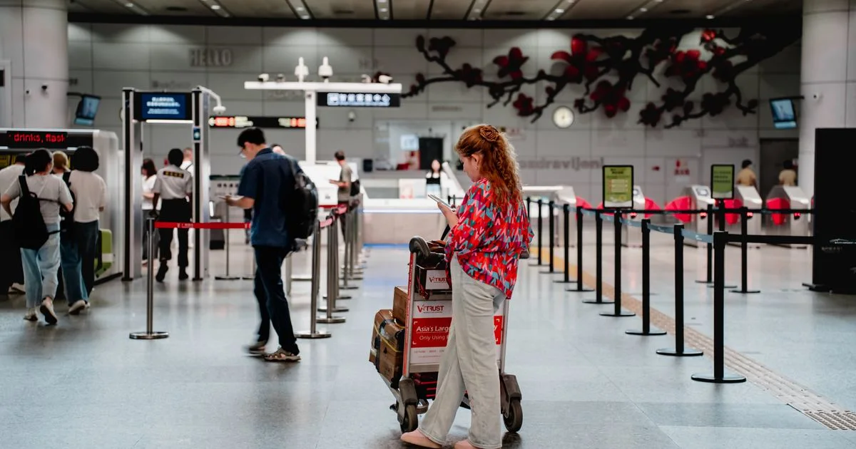 A business traveller checks her phone beside a luggage cart in a bright, modern airport terminal.