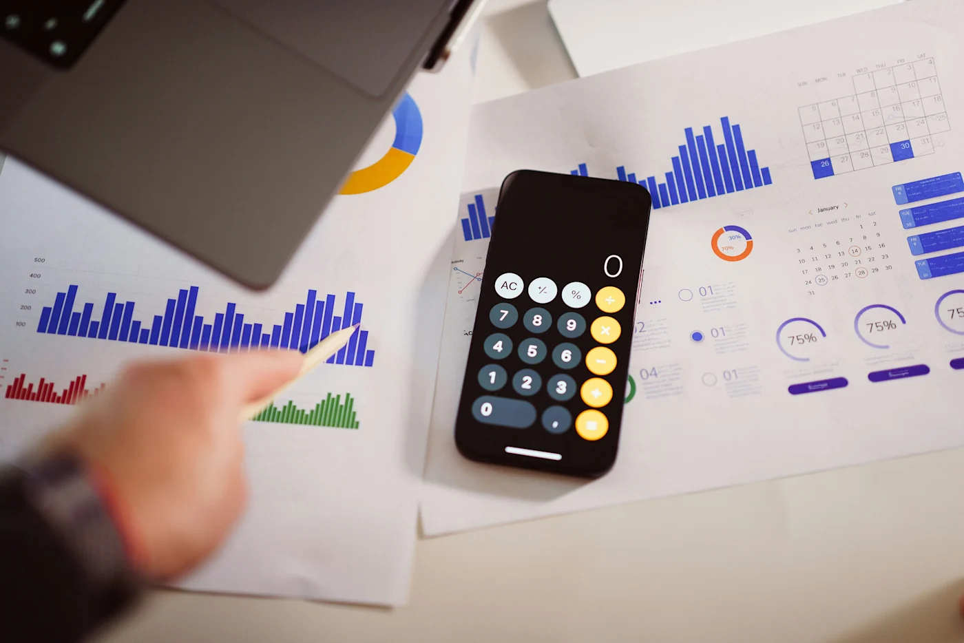 Smartphone calculator resting on printed bar charts beside a laptop and a hand holding a pen
