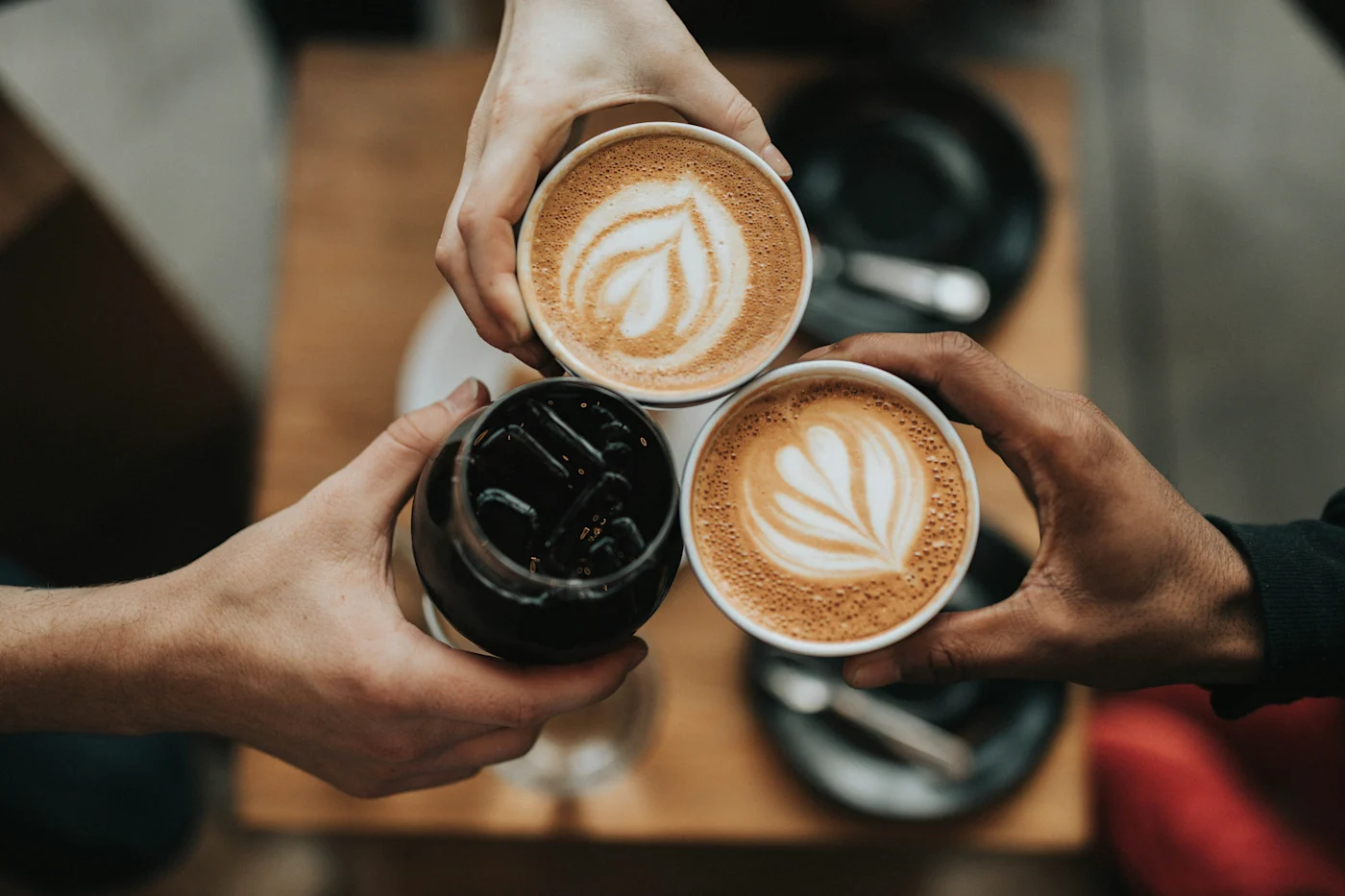 Three hands raising two latte-art coffees and an iced coffee over a wooden cafe table