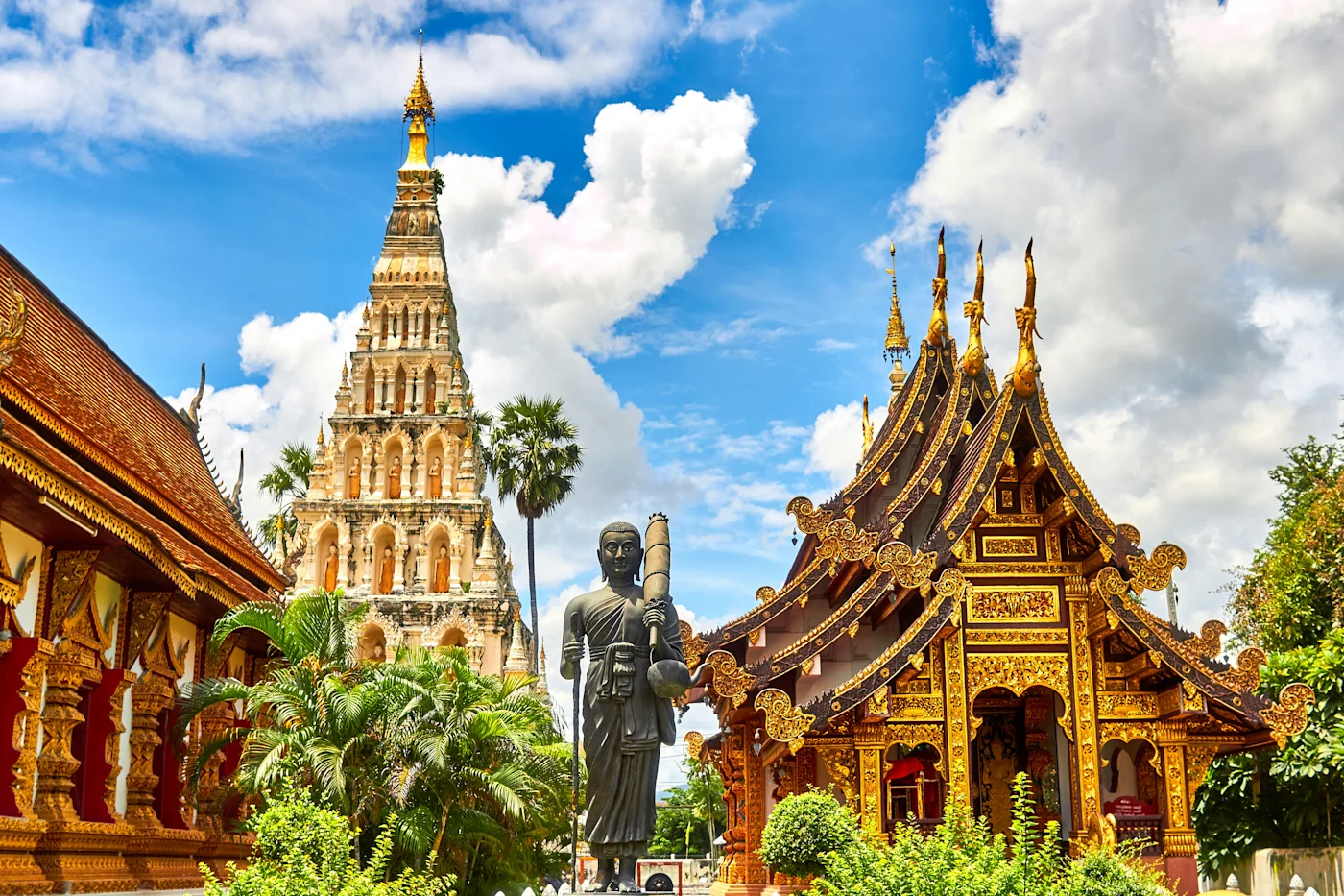 Golden-roofed Thai temple buildings and a brick chedi beside a standing Buddha under blue sky