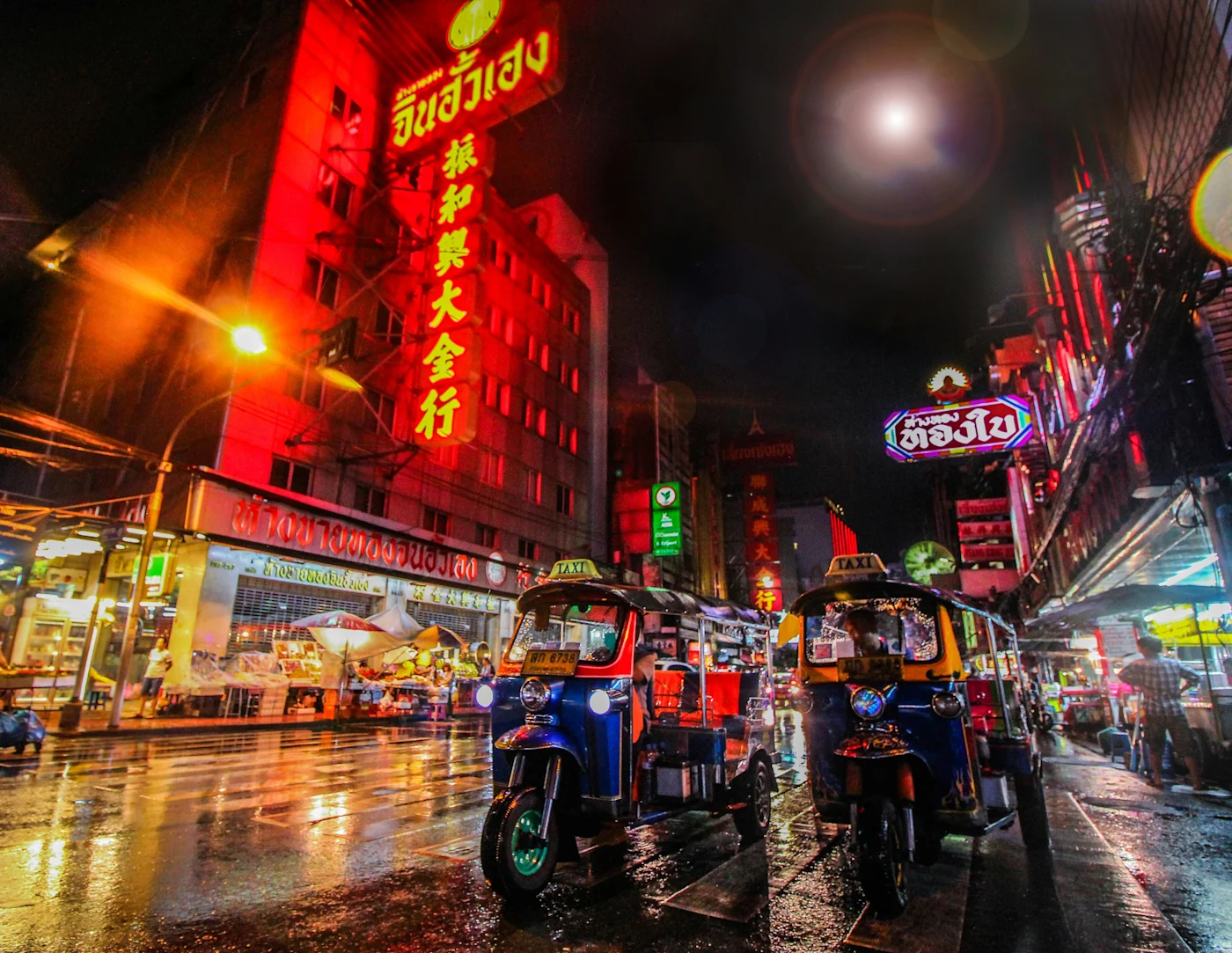 Tuk-tuks parked on a rain-slicked Bangkok Chinatown street glowing with red neon signs