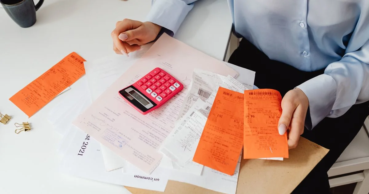Hands sorting paper receipts beside a calculator and notepad on a desk while reviewing travel spend.  (source: https://images.pexels.com/photos/7680736/pexels-photo-7680736.jpeg)