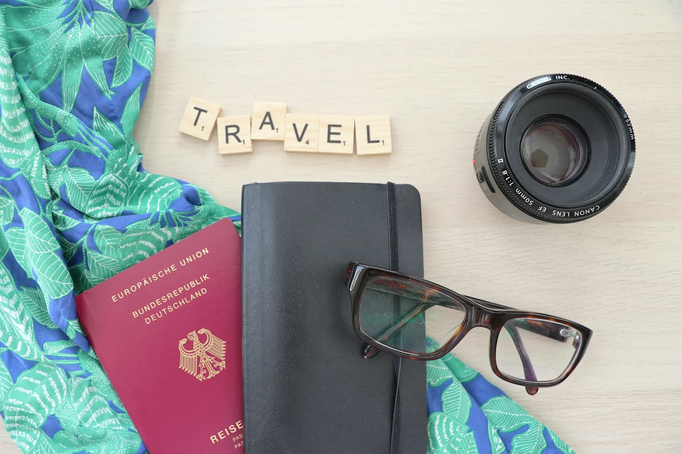 Flat lay of a German passport, notebook, glasses, camera lens and scarf with Scrabble tiles spelling TRAVEL
