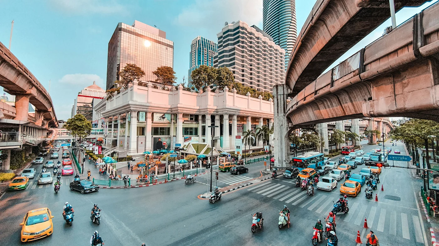Busy Bangkok intersection with taxis and motorbikes below curving Skytrain tracks and high-rise towers