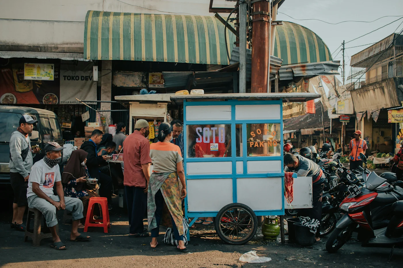 People gathered around a blue street-food cart selling soto beside parked motorbikes and market shops