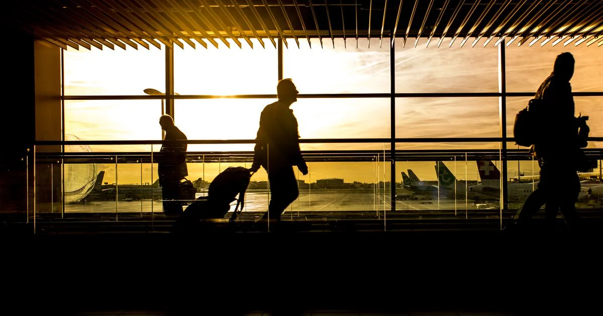 Travellers wheel suitcases through a sunlit airport terminal, silhouetted against a warm golden sunset.