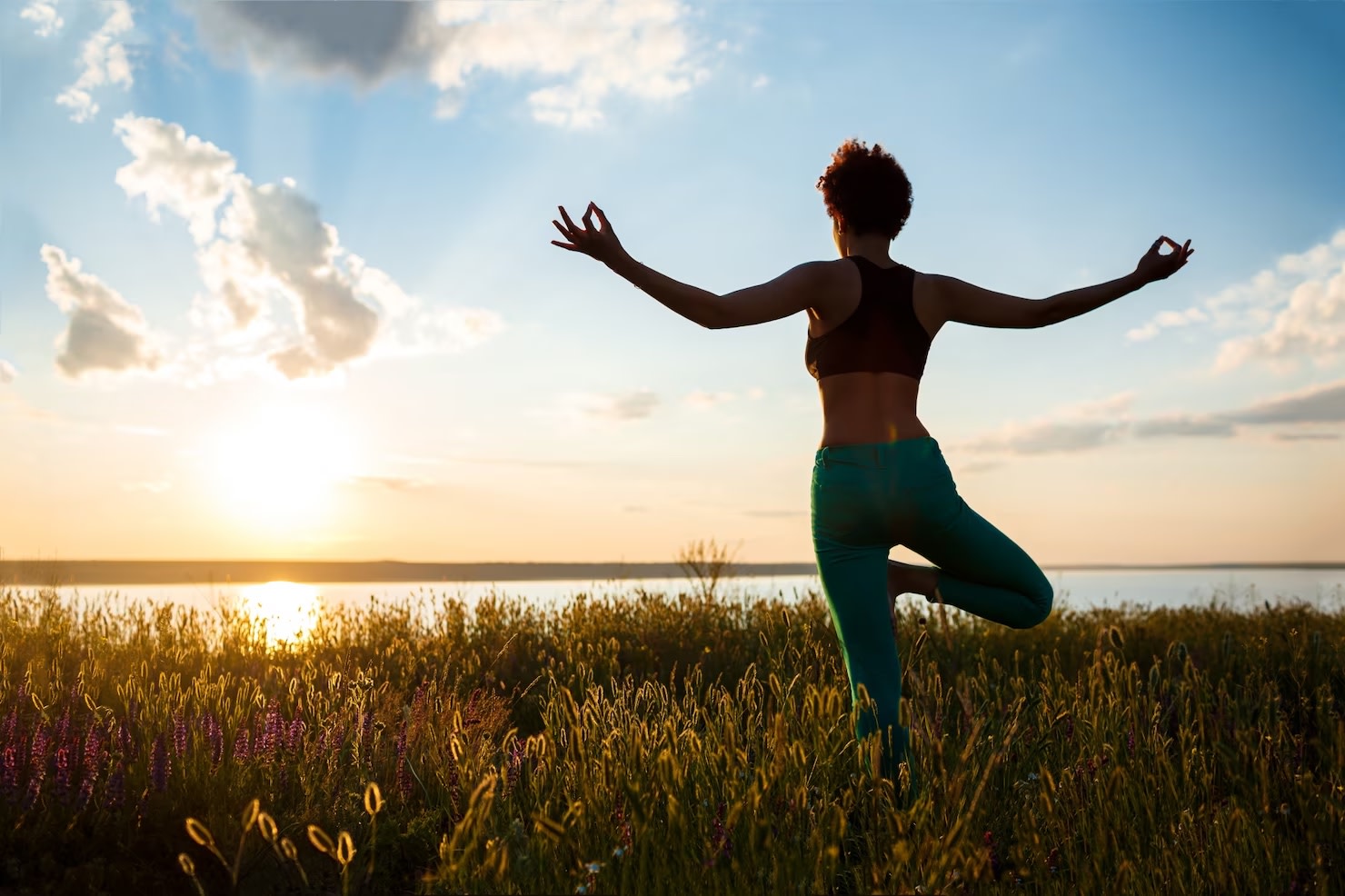 Image of woman standing in field overlooking water while doing yoga pose with sunrise