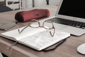 glasses on desk with notebook and laptop open