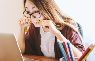 woman biting on pen whilst typing in front of laptop