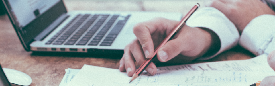 man holding pen whilst writing on paper with laptop besides him