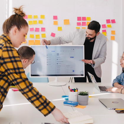 Group of co-workers collaborating around a computer monitor. Sticky notes on the wall behind them.