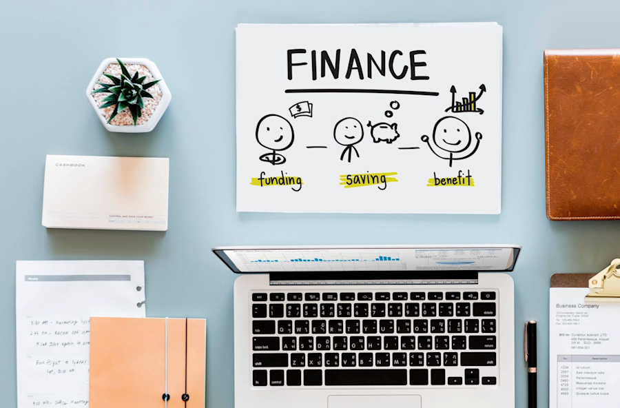 Blue background, computer, pen and paper around and the word finance written on top.