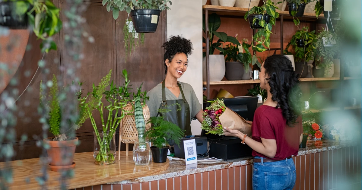 Happy black woman entrepreneur standing behind counter in plant store selling fresh flowers to client
