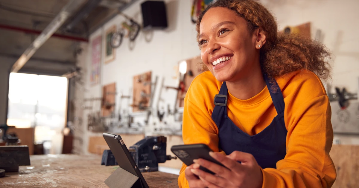 Female Business Owner In Workshop Using Digital Tablet And Holding Mobile Phone