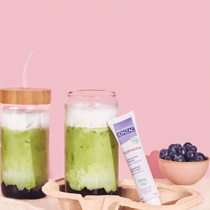 Boisson Blueberry Matcha au collagène à côté d'un tube de crème riche lissante Sublimactive Eau Thermale Jonzac et d'un bol de myrtilles sur fond rose.