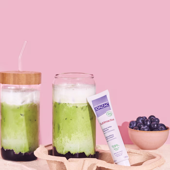 Boisson Blueberry Matcha au collagène à côté d'un tube de crème riche lissante Sublimactive Eau Thermale Jonzac et d'un bol de myrtilles sur fond rose.