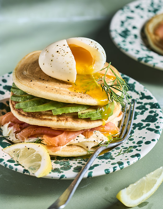 Pile de pancakes salés au saumon fumé, tranches d'avocat frais et crème à l'aneth, surmontée d'un œuf mollet coulant fendu en deux, servis sur une assiette verte mouchetée avec des quartiers de citron.