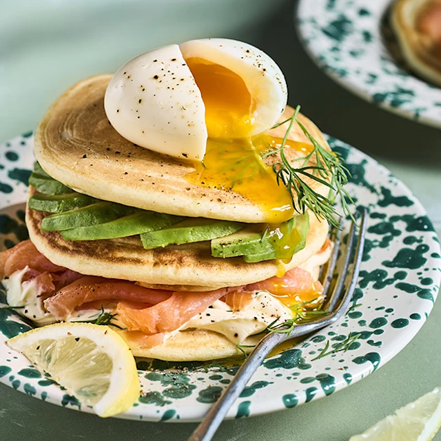 Pile de pancakes salés au saumon fumé, tranches d'avocat frais et crème à l'aneth, surmontée d'un œuf mollet coulant fendu en deux, servis sur une assiette verte mouchetée avec des quartiers de citron.