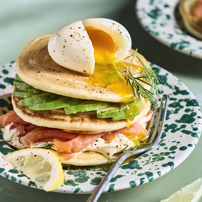 Pile de pancakes salés au saumon fumé, tranches d'avocat frais et crème à l'aneth, surmontée d'un œuf mollet coulant fendu en deux, servis sur une assiette verte mouchetée avec des quartiers de citron.