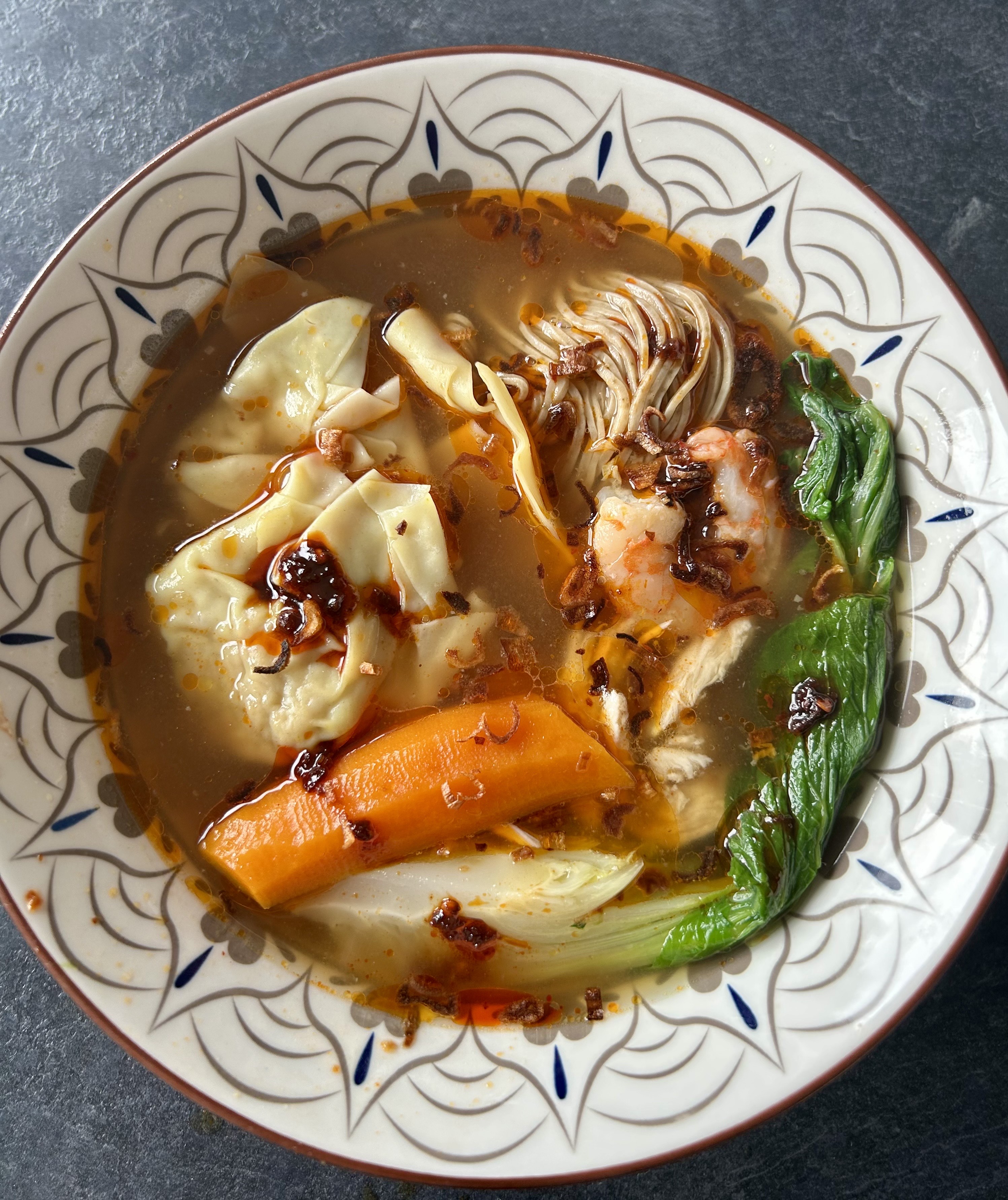 Vue plongeante sur un bol de soupe de wontons maison au poulet et crevettes, recette de @celine.nguyen. On distingue les raviolis, des tranches de carottes, du pak choï, des nouilles et de la chili oil flottant sur le bouillon IBO.