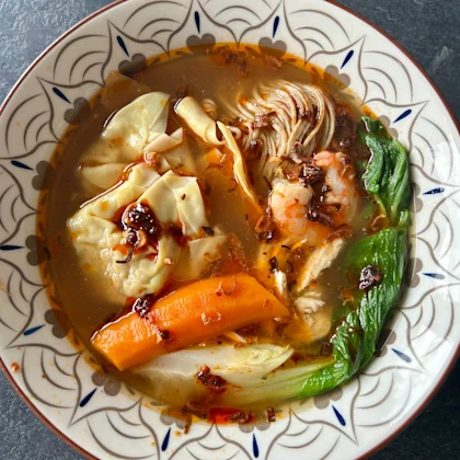 Vue plongeante sur un bol de soupe de wontons maison au poulet et crevettes, recette de @celine.nguyen. On distingue les raviolis, des tranches de carottes, du pak choï, des nouilles et de la chili oil flottant sur le bouillon IBO.