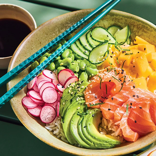 Poke bowl au saumon frais mariné, avocat, mangue, radis, edamames et concombre servi avec des baguettes bleues et une coupelle de sauce soja.