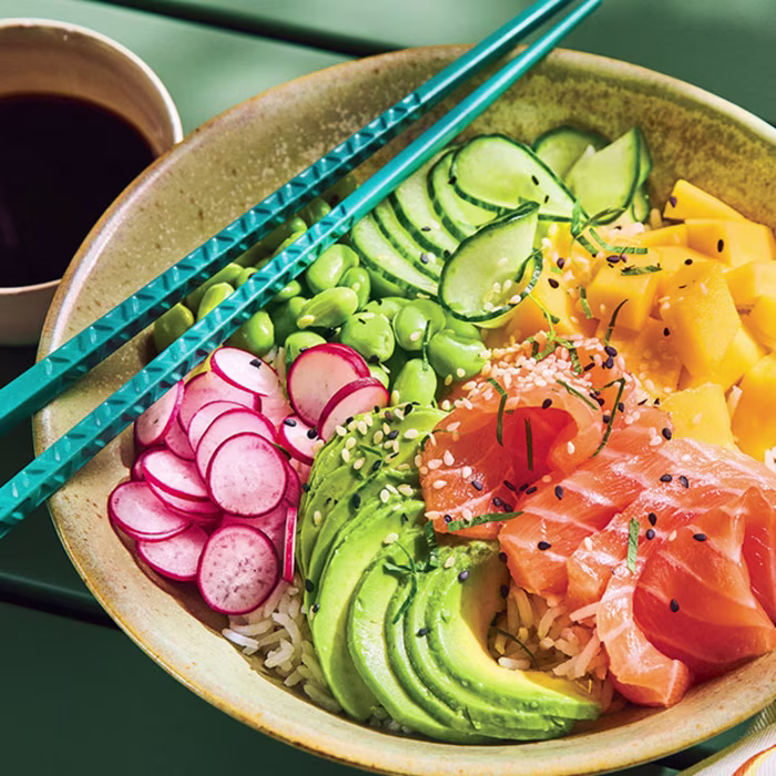 Poke bowl au saumon frais mariné, avocat, mangue, radis, edamames et concombre servi avec des baguettes bleues et une coupelle de sauce soja.
