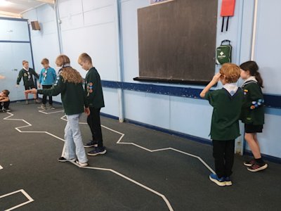 Two Cubs walk blindfolded along a taped zigzag line, given directions by another Cub who can see the line. Other Cubs are waiting their turn at the end of the taped line.