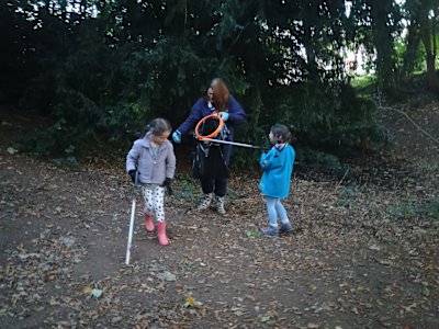 Two young Beavers carrying litter pickers collect rubbish in a wooded area. They are accompanied by an adult volunteer carrying the bag of rubbish, as one of the Beavers raises her picker to drop an item into the bag while the other looks intently on the ground.