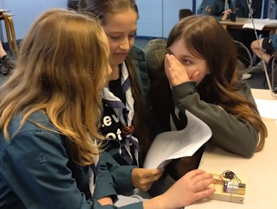 Three Scout girls are grouped around a homemade Morse key, one has her finger on the key, while the other talks furtively behind her hand and the third makes a grimace, while looking at the paper in her hand.