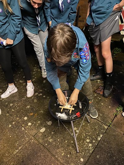 Several Scouts look on as another attempts to get a fire started during the fire lighting race. He's chosen to build his sticks into a log cabin, in the hopes that it helps his fire start quickly.