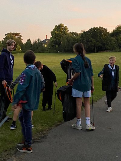 A group of Scouts are picking litter in their local park as the sun sets. One carries a bag holder and a litter picker while the rest carry their own litter pickers.