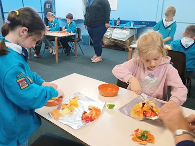 Two Beavers cut up an assortment of fruit. One is standing while the other sits, both are concentrating on their task.