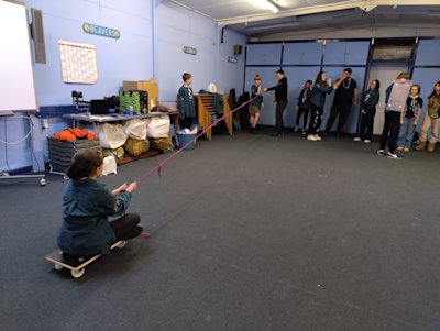 The Scouts are pulling their teammate along the length of the room on a cart, having turned their short rope length into one long length.