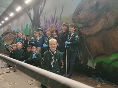 A group of Cubs holding litter pickers are standing and crouching in front of a new mural of wildlife and plantlife along the underpass. Most Cubs are smiling and some are making silly faces.