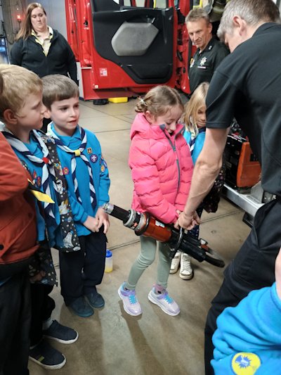 A young Beaver in a bright pink puffy jacket tries to lift a fireman's cutter, assisted by a fireman. It looks heavy! Three other Beavers, their leader and a firefighter look on.