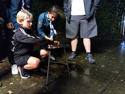 A Scout crouches next to the firebowl to get his fire started, while others keep an eye on his progress.