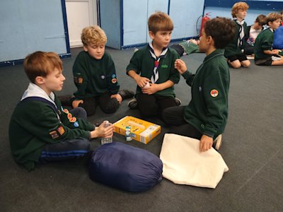 Four Cubs sit on the floor, a small pile of items gathered in the middle of them, that they've chosen from the centre of the room. So far they've chosen a bottle of water, a sleeping bag and a cardboard box. What will they choose next?