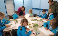 8 Beaver Scouts are sat around a cluster of tables, which are covered in leaves, sheets of paper and thick crayons. The Beavers are busy making leaf rubbings. They are being assisted by an adult and a teenager.