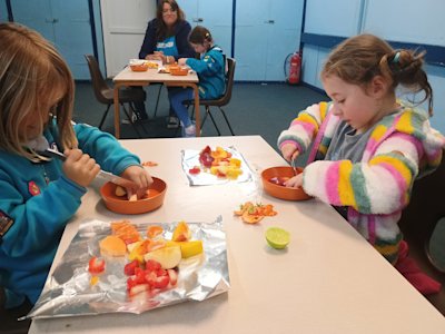 Two Beavers have begun cutting up their strawberries, melon, pineapple, mango and apple to create their fruit salad. A pile of chopped fruit is on a piece of foil while they chop more fruit in a shallow, orange melamine bowl.