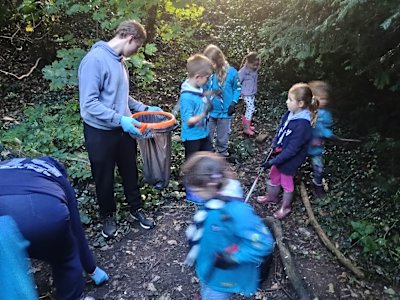 A group of Beavers, litter pickers in hand, are collecting rubbish in a woodland setting. A teenage volunteer holds a bag for them to drop their trash into as they find it.