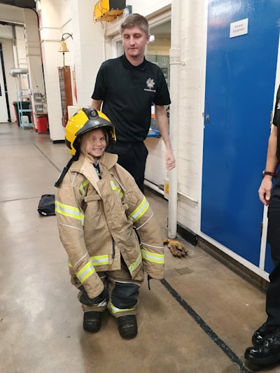 A Beaver stands in a pair of fireman's boots which are enormous on her, wearing a fireman's trousers and coat that are equally huge on her. The fireman standing behind her has just put the fireman's helmet on her head, and she has a big cheeky grin on her face.