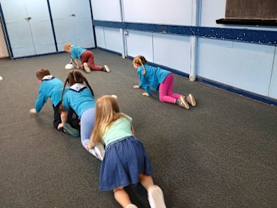 Three Beavers are crawling down the hall, holding onto each other's ankles in a chain, racing the other team. The second team has broken apart and the two Beavers on that team are crawling with a large space between them, so they will not win.