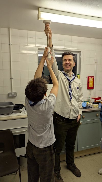 With no hanging bulbs in the Scout hut, one of the taller leaders is holding the bulb bracket as high as he can, so the Scout can attempt to change the lightbulb above his head.