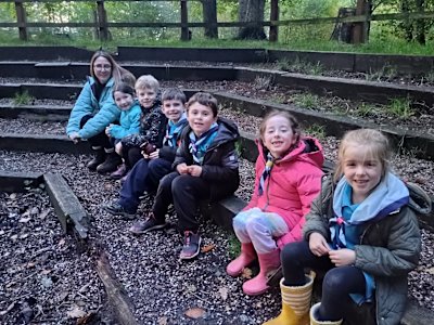 Six Beavers sit with an adult helper, smiles on their faces, waiting for the campfire singing to begin once the other groups arrive.