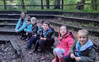 Six Beavers sit with an adult helper, smiles on their faces, waiting for the campfire singing to begin once the other groups arrive.