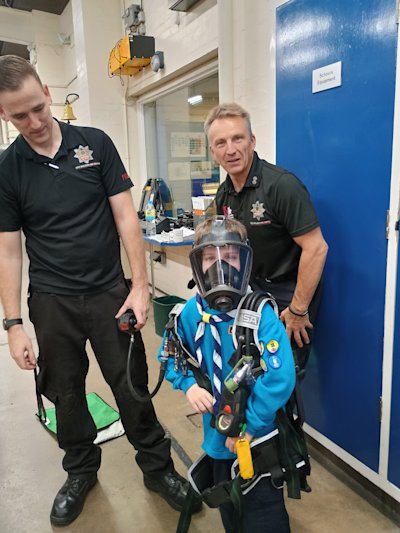 One of the Beavers is wearing a firefighter's breathing apparatus, without the tank. Two firefighters stand next to him, having helped him put on the gear.