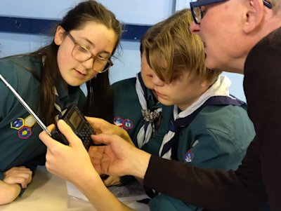 A Scout holds a small radio in his hands while two other Scouts look on. A local expert is showing the Scouts how to use the radio.