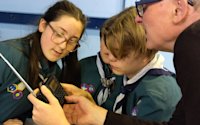 A Scout holds a small radio in his hands while two other Scouts look on. A local expert is showing the Scouts how to use the radio.